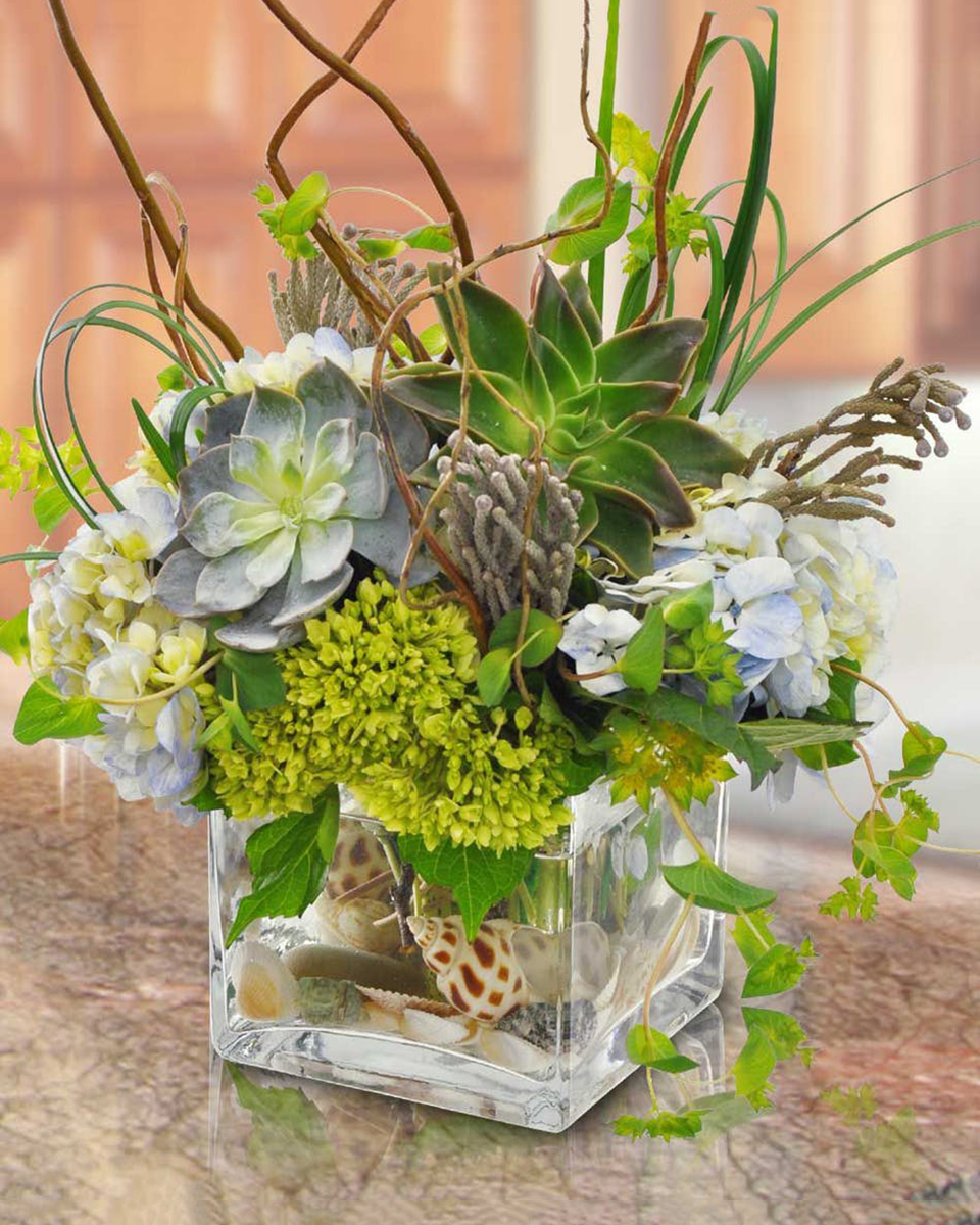 Cool and Breezy Standard (5 x 5 in cube) A 5 x 5 Glass Cube is overflowing with succulents, hydrangea, curley willow, and exotic greenery and is anchored with sea shells and rocks.
DELIVERY: Every order is hand-delivered direct to the recipient. These items will be delivered by us locally, or a qualified retail local florist.
