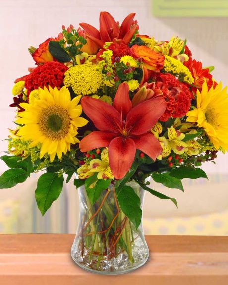 Autumn Equinox-A Fall feast of flowers. Red tiger lilies, yellow sunflowers, yellow alstroemaria, yellow yarrow,red hypericom, yellow dot poms, and seasonal greens are designed in a clear bunch vase that is decoratively lined with curley willow.-Fall Arra